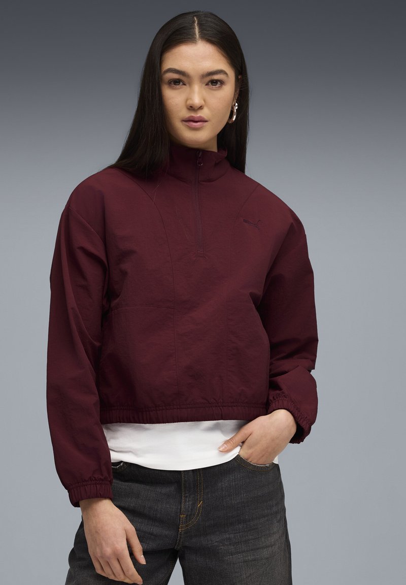 Ung kvinde med langt mørkt hår iført en marsala-farvet zip-up jakke over en hvid skjorte og mørke jeans, der står med den ene hånd i lommen.