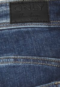 Tecido de denim azul escuro com linhas de costura visíveis e um rótulo de couro preto marcado "ONLY" na cintura.