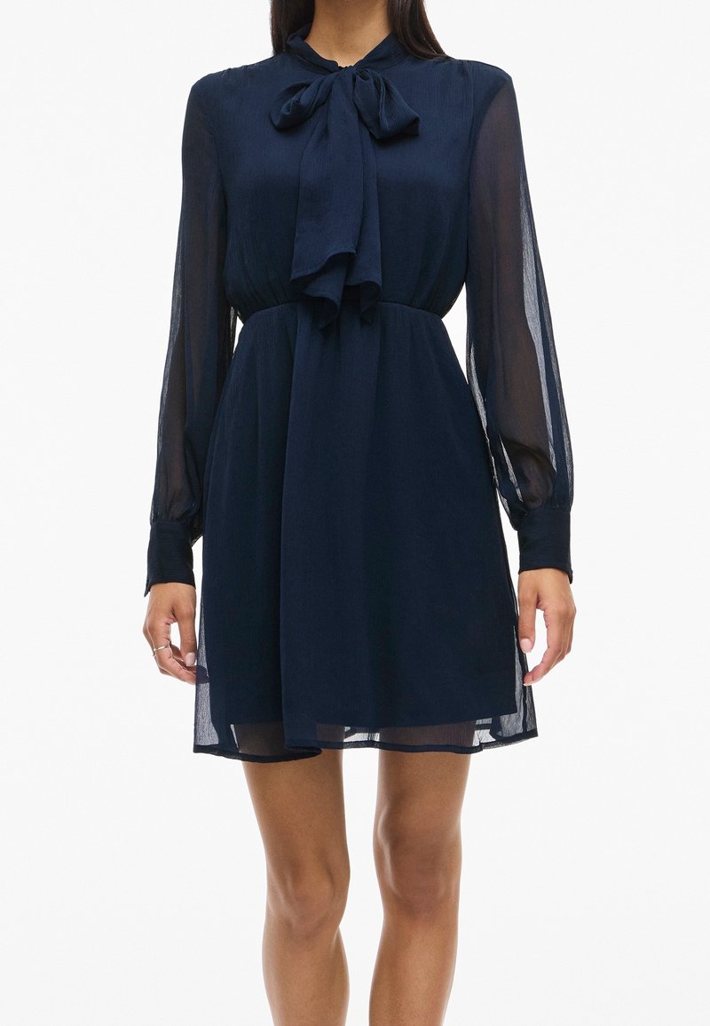 Femme portant une robe bleu marine à manches longues avec des manches transparentes et un détail nœud papillon au col, debout devant un fond uni.