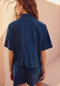 Mörk denimskjorta med kort ärm och boxig passform, med rak fåll och synliga sömdetaljer, bärs med matchande shorts.