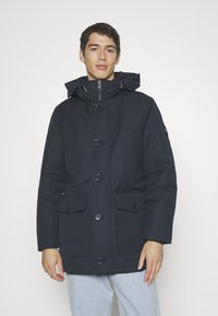 Navy blue parka jacket with a hood, featuring large front pockets, button closure, and a smooth texture. Classic casual design.