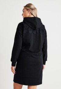 Črna kapuča obleka s čipkastim detajlom na ramah, dolgimi rokavi in sproščenim krojem, ki seže do sredine stegna.