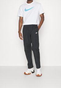 Vit t-shirt med en blå Nike-logotyp, matchad med svarta cargobyxor med elastisk midja och sidofickor samt en liten vit logotyp. Vita och orange sneakers.