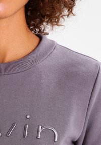 Sudadera gris con cuello acanalado, que presenta un logo metálico bordado de "Calvin" en el frente. Textura suave y diseño casual.