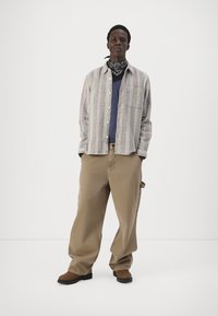 Grå og hvit stripete skjorte med knapper over en marineblå t-skjorte, kombinert med løse beige bukser og brune støvler. Sort bandana rundt halsen.