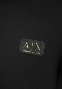 Juodas medvilninis marškinėliai su stačiakampiu juodu odiniu pleistru, ant kurio yra auksu įspausti tekstai "A/X" ir "ARMANI EXCHANGE". Ilgomis rankovėmis.