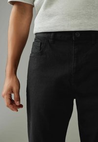 Pantaloni din denim negru cu o croială normală, având un singur buzunar frontal, bretele pentru curea și închidere cu buton; textură netedă și design curat.