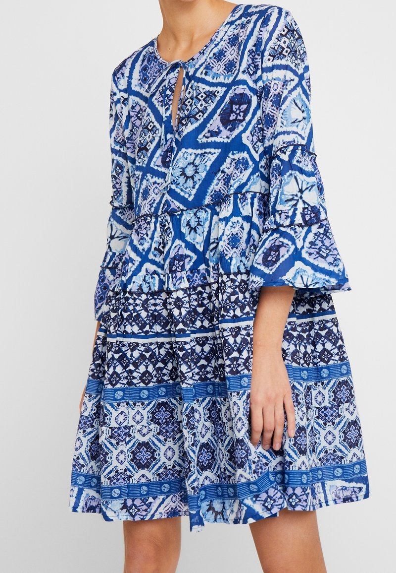Modrá šaty se vzorem, s výstřihem ve tvaru klíčové dírky a rozšířenými rukávy. Obsahují vrstvený, volánkový design s různými odstíny modré a geometrickými vzory.