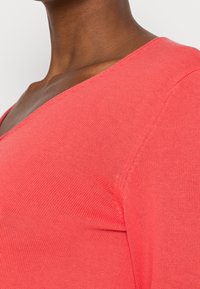 Haut tricot corail avec un design en V. Le tissu présente une fine texture côtelée, mettant en valeur une coupe ajustée et une finition lisse.