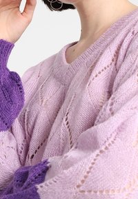 Ljuslila stickad tröja med vid halsringning, försedd med ett öppet diamantmönster och lila ribbade manschetter. Mjuk textur och avslappnad passform.