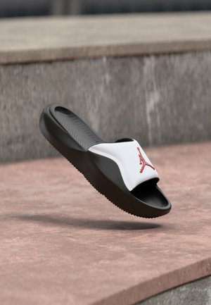 Ciabatta slide bianca e nera con logo Jumpman rosso, che fluttua sopra una superficie di cemento rosa e grigia.