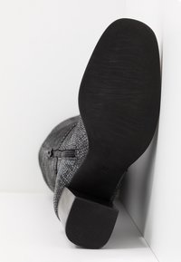 Botte avec motif texturé en peau de serpent grise, fermeture éclair latérale, semelle et talon épais noirs, vue de la semelle sur fond blanc.