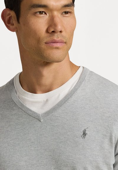 Suéter de malha cinza claro em V, sobre uma camisa branca, apresentando um pequeno logotipo escuro no lado esquerdo do peito. Textura suave, design casual.