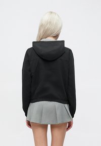 Μαύρη κουκούλα με cropped σχέδιο, ελαστική μέση και λεία υφή, σε συνδυασμό με μια πλισέ γκρι φούστα.