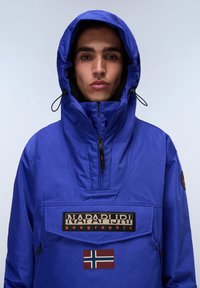 Blue insulated jacket with a high collar and adjustable hood. Features a front pocket, branded patch, and small Norwegian flag detail.