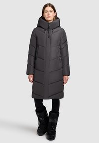 Lång, mörkgrå puffjacka med hög krage, dragkedja fram och quiltat mönster. Modellen har på sig svarta stövlar och leggings.