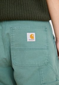 Mintgröna cargobyxor med en bakficka med en vit etikett och en gul logotyp. Texturerad olivgrön tröja syns delvis ovanför.