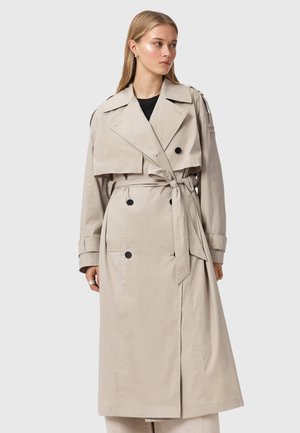 Vrouw met een beige dubbelgeknoopte trenchcoat met zwarte knopen en een gestrikte tailleband, kijkt opzij tegen een effen achtergrond.