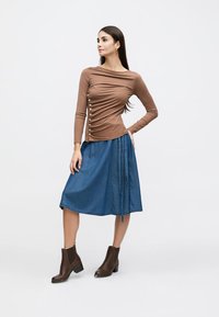 Γυναίκα που στέκεται με το χέρι στη μέση, φορώντας καφέ μακρυμάνικη μπλούζα με κουμπιά στο πλάι, μπλε μίντι φούστα και καφέ μποτάκια στον αστράγαλο, πάνω σε λευκό φόντο.