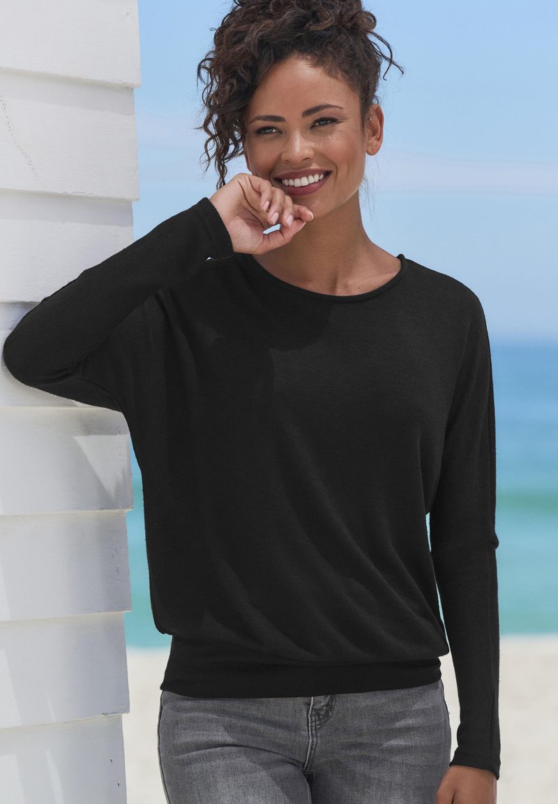 Schwarzes Langarmshirt mit lockerer Passform und rundem Halsausschnitt, kombiniert mit hellgrauen Jeans, vor einem Strandhintergrund.