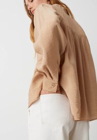 Camicia beige a maniche lunghe con tessuto testurizzato e polsini abbottonati, abbinata a jeans bianchi con tasca posteriore e cuciture semplici.