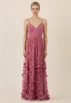 Frau steht aufrecht und trägt ein bodenlanges, staubrosafarbenes Kleid mit dünnen Trägern und floralen Applikationen am Rock.