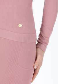 Maglietta a maniche lunghe realizzata in tessuto a coste di un delicato rosa chiaro, con un emblema decorativo dorato vicino all'orlo e un dettaglio di tasca laterale visibile.