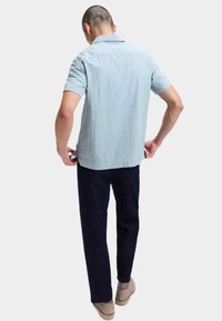 Man in een lichtblauw, gestructureerd, kortarmig shirt, donkere broek, beige schoenen, staand met de handen die de zoom van het shirt aanpassen, met de rug naar de kijker.