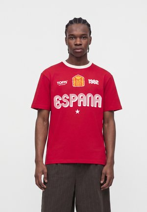 Giovane uomo con capelli intrecciati che indossa una maglietta rossa "España" con uno stemma della Spagna, il logo "TOFFS" e la scritta "1982", in piedi su sfondo bianco.