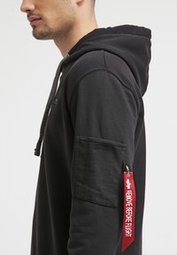 Sudadera negra con cordones, que cuenta con un bolsillo de parche texturizado en la manga y una etiqueta roja de "RETIRE ANTES DEL VUELO".
