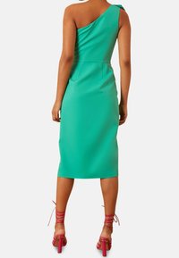 Robe ajustée à une épaule, de couleur vert vif, longueur genou, en tissu lisse. Associée à des sandales à talons hauts roses avec des lanières.