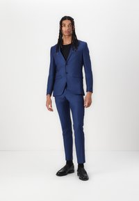 Costume sur mesure bleu avec deux boutons, revers en dents de scie et pantalon slim. Chaussures noires et chemise portée en dessous. Texture de tissu lisse.
