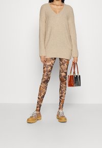 Beżowy sweter dzianinowy z dekoltem w serek, zestawiony z brązowymi legginsami w kwiatowy wzór. Modelka trzyma małą, fakturowaną torebkę i nosi brązowe, masywne buty.