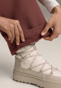 Ruce upravují manžetu žebrovaných bordových kalhot nad béžovou polstrovanou zimní botou s tlustou podrážkou a překříženými tkaničkami na neutrálním pozadí.