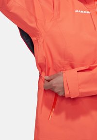 Hand pulling zipper on bright orange Mammut jacket sleeve with Velcro cuff and underarm vent open.