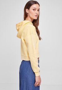 Felpa gialla accorciata in morbido cotone, con cappuccio, spalle scese e polsini a coste, abbinata a pantaloni di jeans a vita alta.