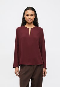 Γυναίκα με σγουρά μαλλιά, φορώντας μια μακριά μανίκι μπορντό μπλούζα με μια μικρή σχισμή και κούμπωμα στον λαιμό και σκούρο καφέ παντελόνι, όρθια.