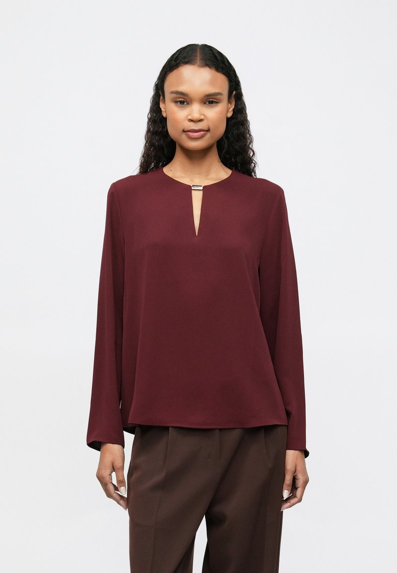 Γυναίκα με σγουρά μαλλιά, φορώντας μια μακριά μανίκι μπορντό μπλούζα με μια μικρή σχισμή και κούμπωμα στον λαιμό και σκούρο καφέ παντελόνι, όρθια.