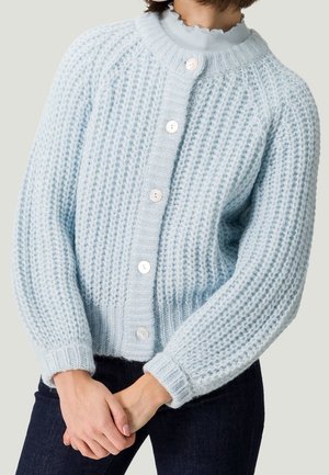 Kobieta w jasnobłękitnym, grubym kardiganie z białymi guzikami i falowanym dekoltem, w ciemnych jeansach, ze splecionymi rękami.