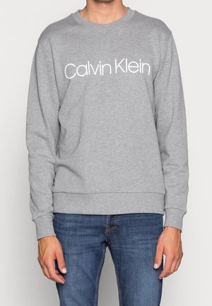 Person, die einen grauen Calvin Klein Pullover und blaue Jeans trägt, steht mit entspannten Händen an den Seiten vor einem einfarbigen Hintergrund.