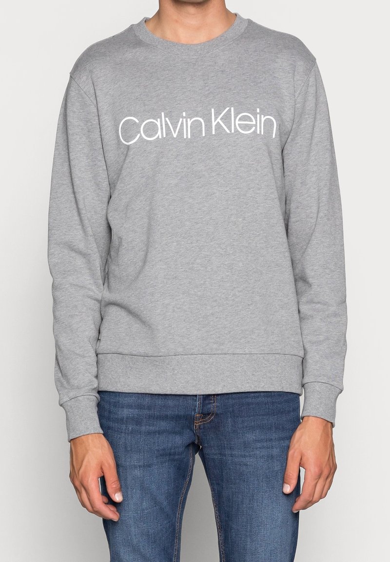 Personne portant un sweat-shirt gris Calvin Klein et un jean bleu, debout avec les mains détendues le long du corps devant un fond uni.