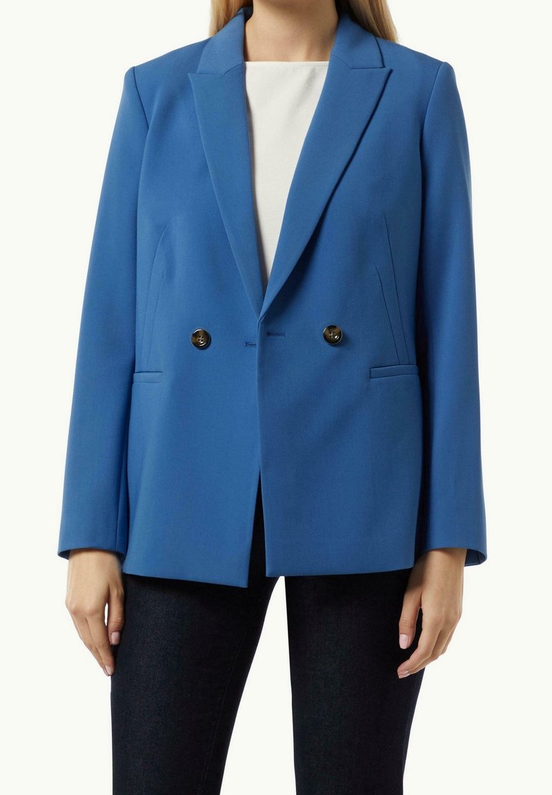 Femme portant un blazer bleu croisé avec des boutons noirs sur un haut blanc et un pantalon noir, debout devant un fond uni.