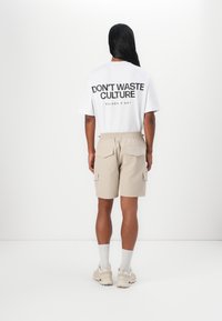 Hvid t-shirt med sort tekst "DON'T WASTE CULTURE" på ryggen, parret med beige shorts med sidelommer og baglommer.