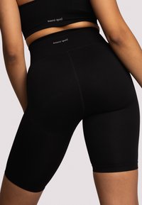 Žena v černých bezešvých vysokých biker kratasech a odpovídajícím krátkém topu s logem "oceans apart" na pásku a na zádech.