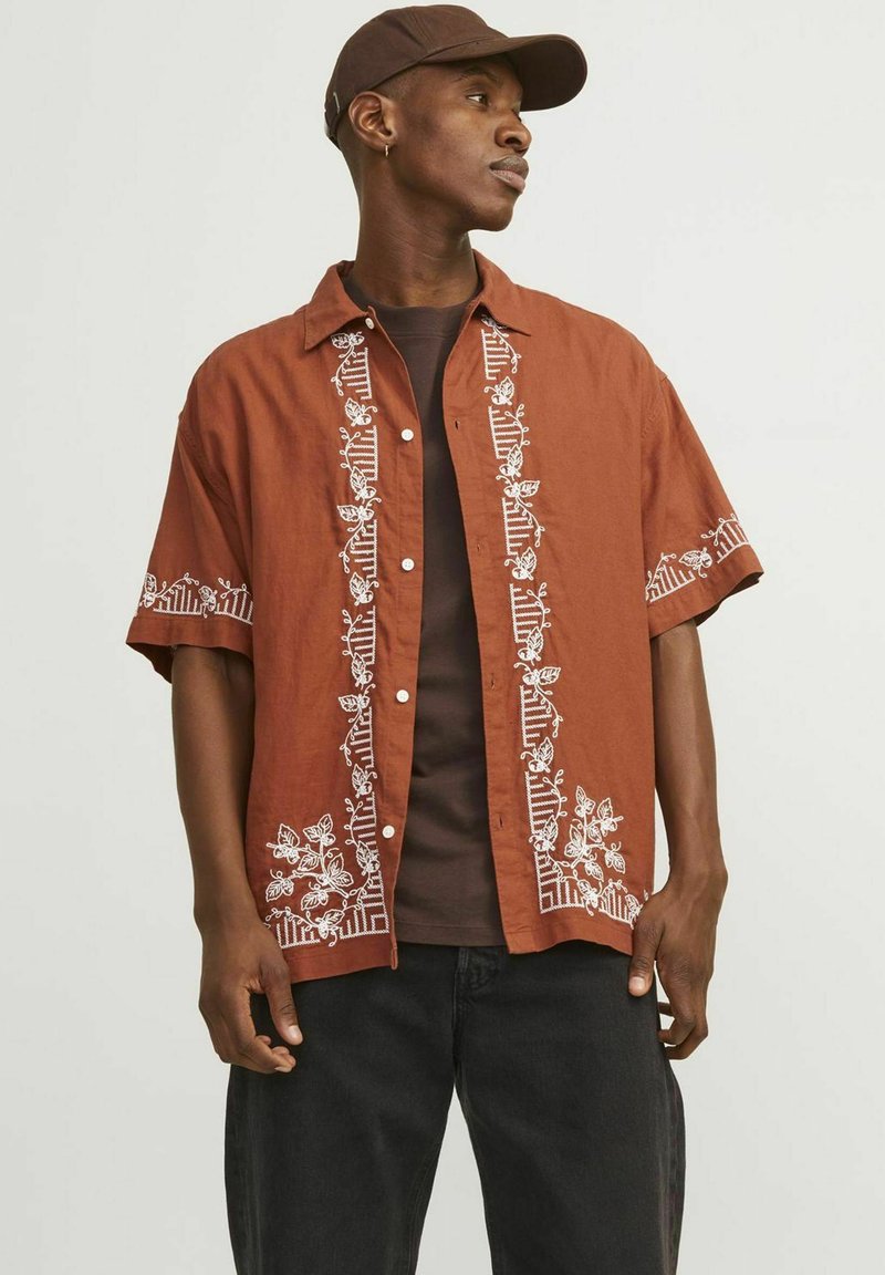 Chemise marron à manches courtes avec une broderie florale blanche le long de la patte de boutonnage et de l'ourlet, associée à une chemise noire et un jean noir.
