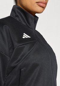 Črna športna jakna z visokim ovratnikom, z zadrgo in belim logotipom na rami. Material izgleda gladek in teksturiran.