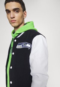 Schwarz-weiße Collegejacke mit einem Seattle Seahawks Logo. Getragen über einem leuchtend grünen Hoodie, der kontrastierende Farben und Texturen zur Schau stellt.