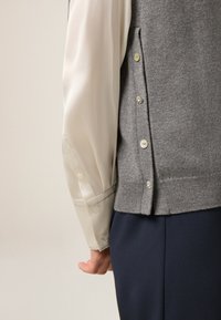 Chaleco gris de punto con botones laterales, usado sobre una camisa blanca de satén de manga larga. Enfoque en las texturas y la tela suave de la camisa.