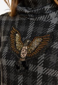 Grau und schwarz karierter Pullover mit einem perlenbestickten Adler-Motiv, das goldene, bronzefarbene und schwarze Akzente aufweist. Weicher Stoff mit hohem Kragen.