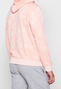 Sudadera con capucha teñida en rosa claro con bolsillo tipo canguro, textura suave, dobladillo y puños acanalados. Combinada con pantalones grises en un ambiente informal.